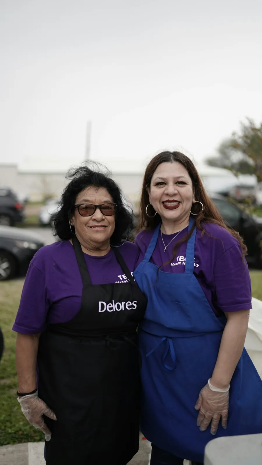 Two volunteers in aprons laughing together at a community food event
